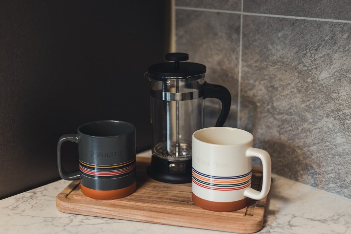 Close-up of a kitchen counter with a French press and two Pendleton mugs on a wooden tray. The countertop is light marble-patterned, and the backsplash is gray tile.