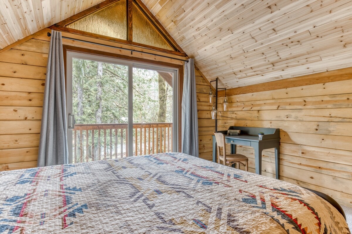 Bedroom with a large sliding glass door leading to a balcony, offering views of lush trees. The room features wood-paneled walls and a sloped ceiling, with a bed covered in a colorful patterned quilt.