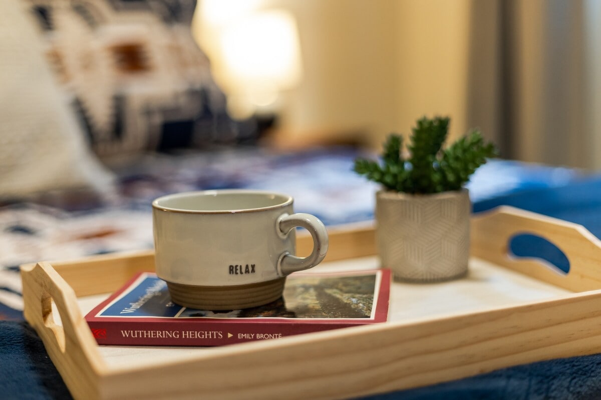 A close-up shot of a wooden tray on the bed, featuring a cream "RELAX" mug, a small potted succulent, and a copy of "Wuthering Heights" on a red book cover.