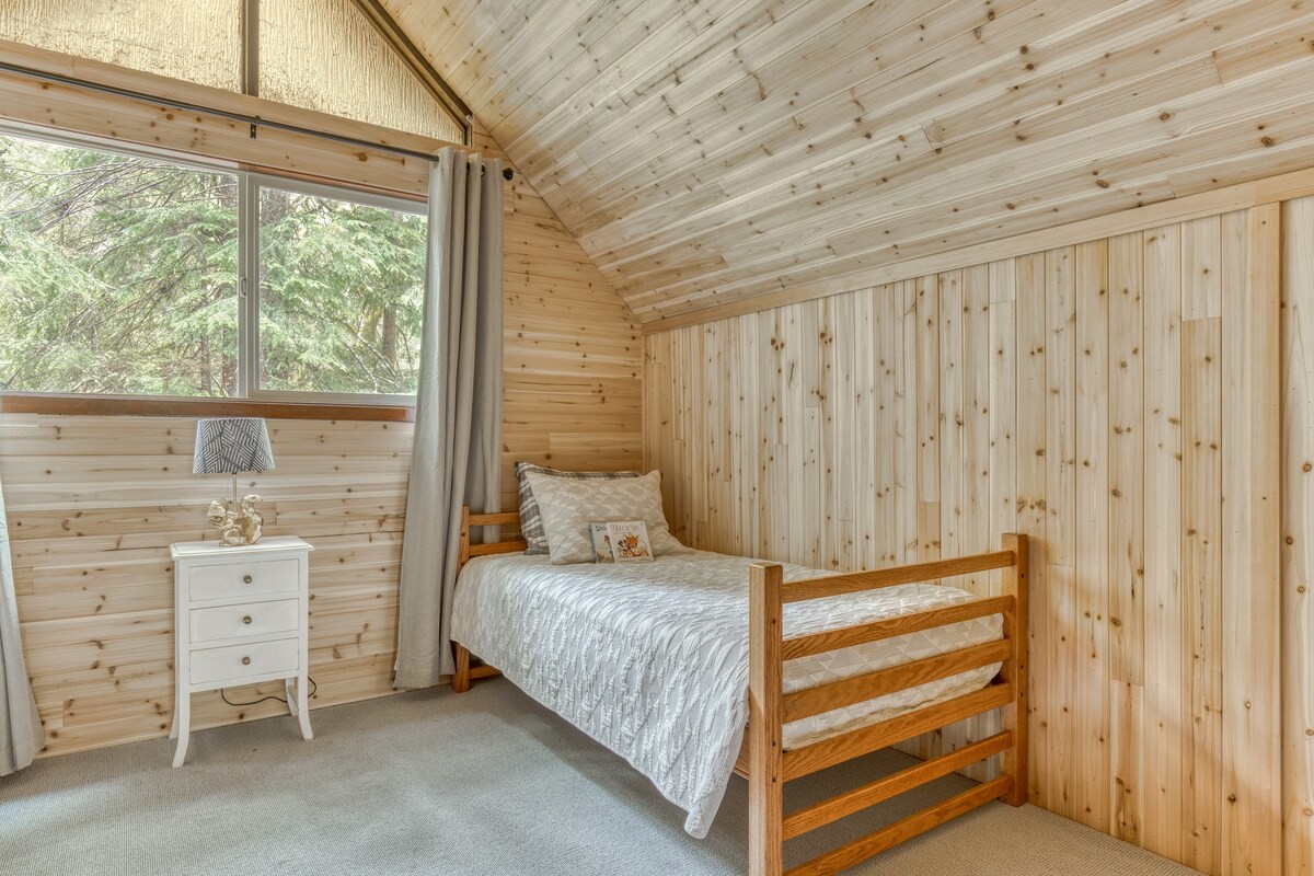A cozy bedroom with light wood paneling on walls and a sloped ceiling, featuring a single bed with white bedding, a small white nightstand with a lamp, and a large window looking out to lush trees.