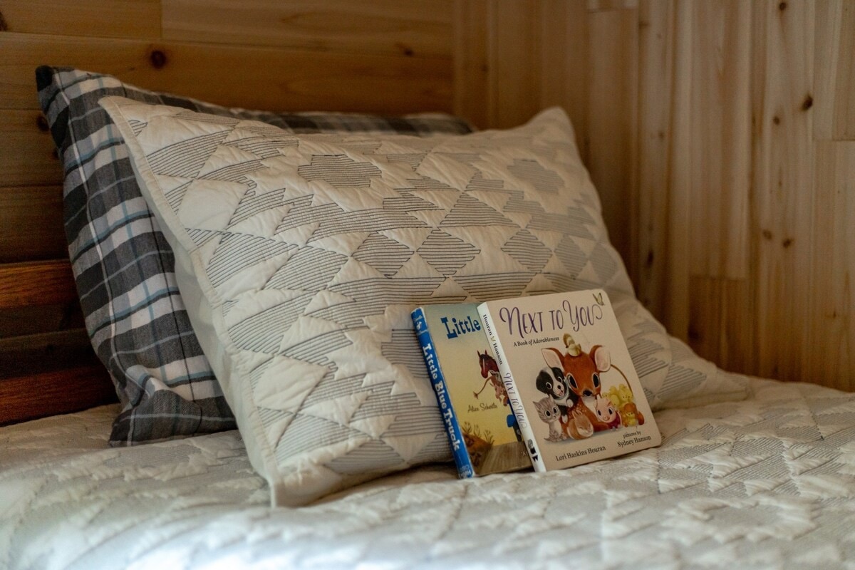 A close-up of a bed with a white quilted comforter and two patterned pillows, displaying two children's books titled 'Little Blue Truck' and 'Next to You'.