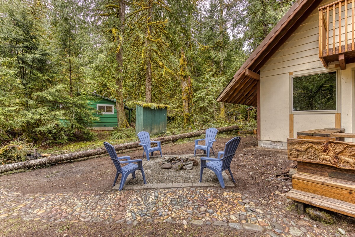 An outdoor fire pit area with four blue Adirondack chairs arranged around a stone fire pit, set on a gravel and stone patio, surrounded by tall evergreen trees and a rustic wooden bench.