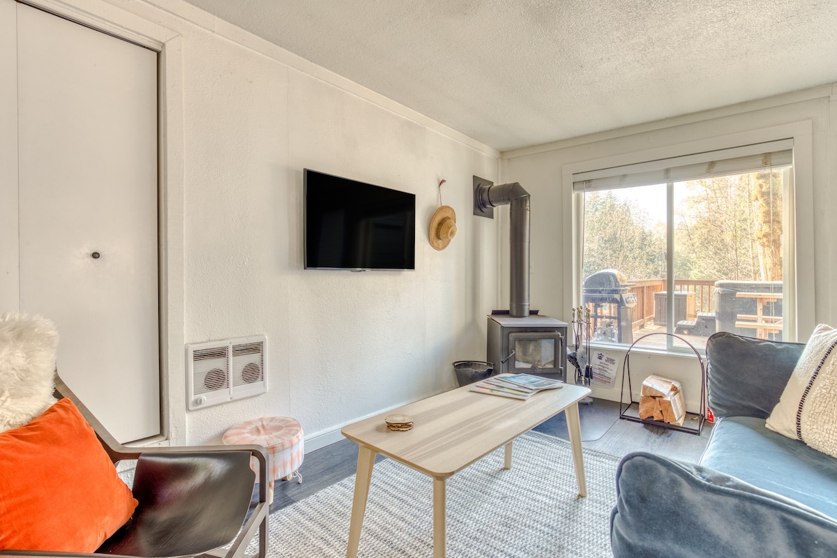 View of the living room from a different angle, showing a wall-mounted TV, a wood-burning stove with a black pipe, and a large window overlooking an outdoor deck. A light-colored coffee table sits on a grey patterned rug, with a modern armchair to the left.