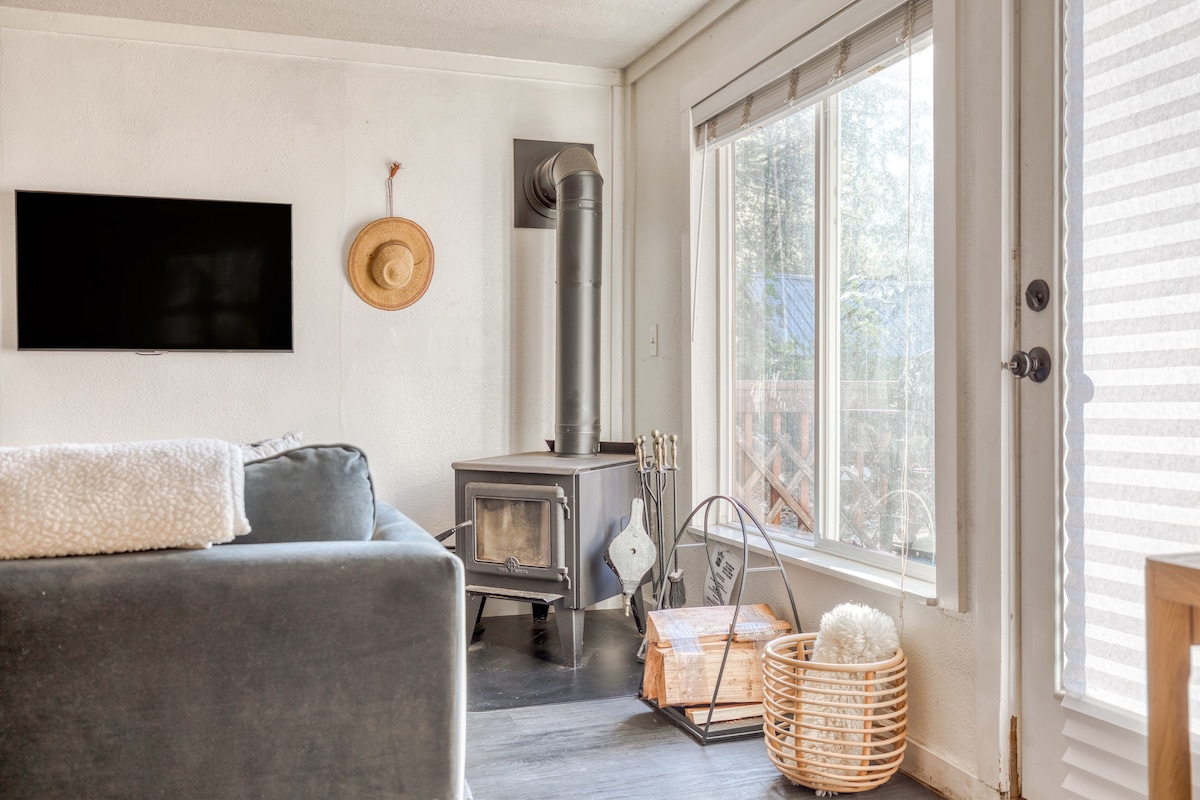 Corner of the living room showing a wood-burning stove with a black pipe, a large window with white blinds, and a glass door leading outside. A light blue sofa is partially visible, along with a basket of firewood and a metal log holder.