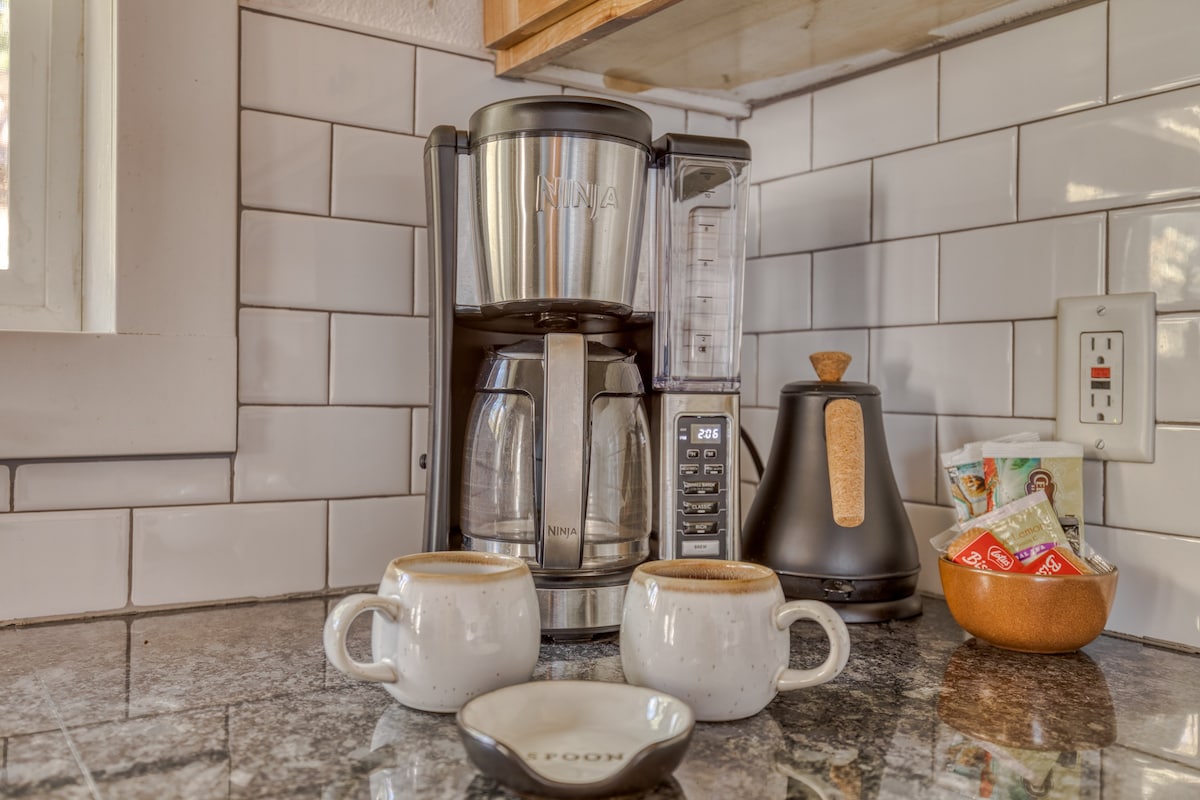 Close-up of a coffee station on a dark granite countertop, featuring a stainless steel Ninja coffee maker, a black electric kettle, two ceramic mugs, and a bowl of coffee pods against a white subway tile backsplash.
