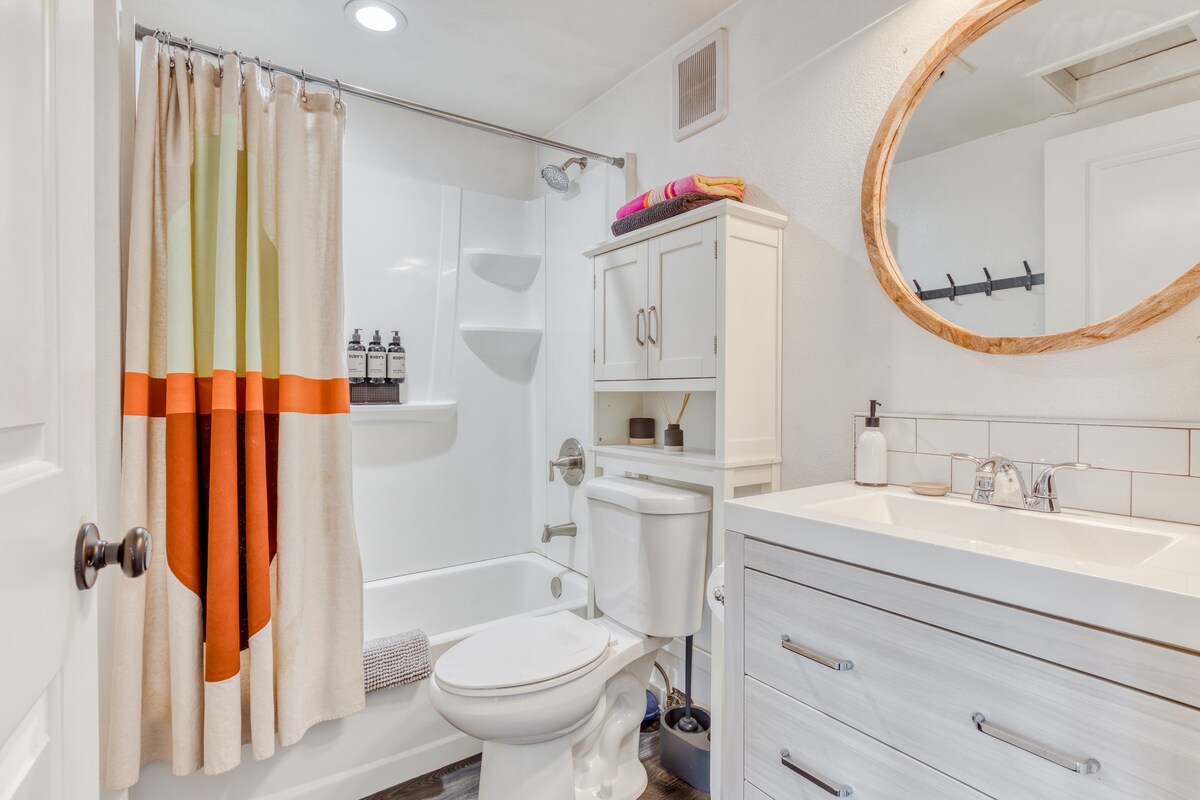 Modern bathroom with a white vanity, round wooden-framed mirror, white subway tile backsplash, and a shower/tub combo with a vibrant striped curtain. A white storage cabinet is positioned above the toilet.