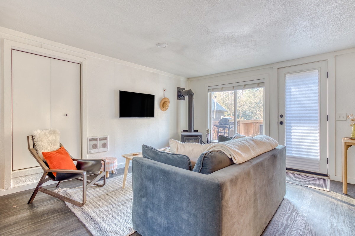 Living room with a light blue velvet sofa facing a wall with a TV and wood-burning stove. A large window and a glass door lead to an outdoor area, with a light-colored coffee table and a grey patterned rug in the foreground.