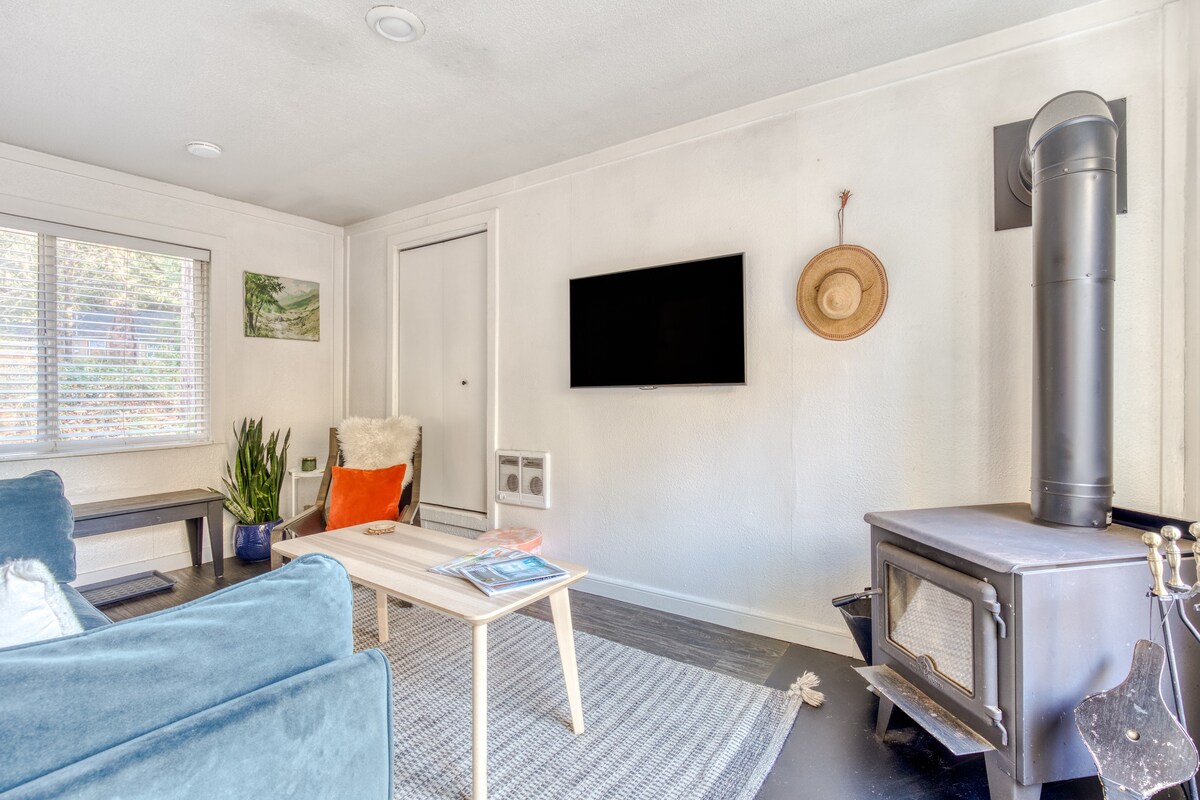 Bright living room with white walls, dark wood-look flooring, and a light blue velvet sofa. A wall-mounted TV, a wood-burning stove, and a window with blinds are visible, along with a light-colored coffee table on a grey patterned rug.