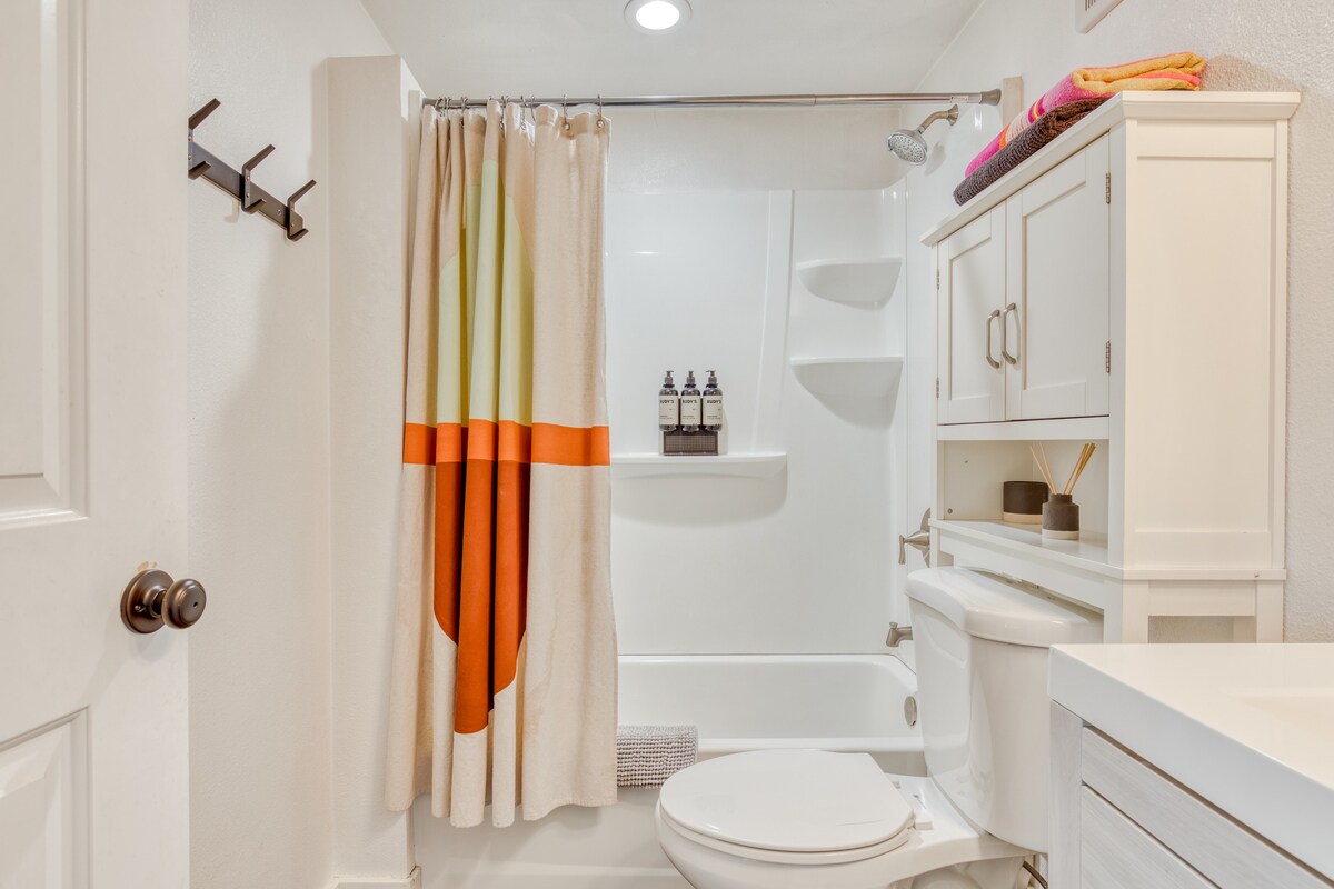 Close-up view of the bathroom's shower/tub combo with a colorful striped curtain, a white toilet, and a tall white storage cabinet. Shower amenities are neatly arranged on a shelf within the shower.