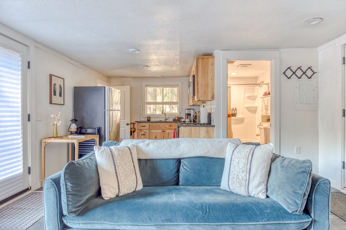 Front view of the light blue velvet sofa with white throw pillows and a blanket, facing away from the camera. The background shows a partial view of the kitchen with wooden cabinets and a stainless steel refrigerator, and an open doorway to the bathroom.