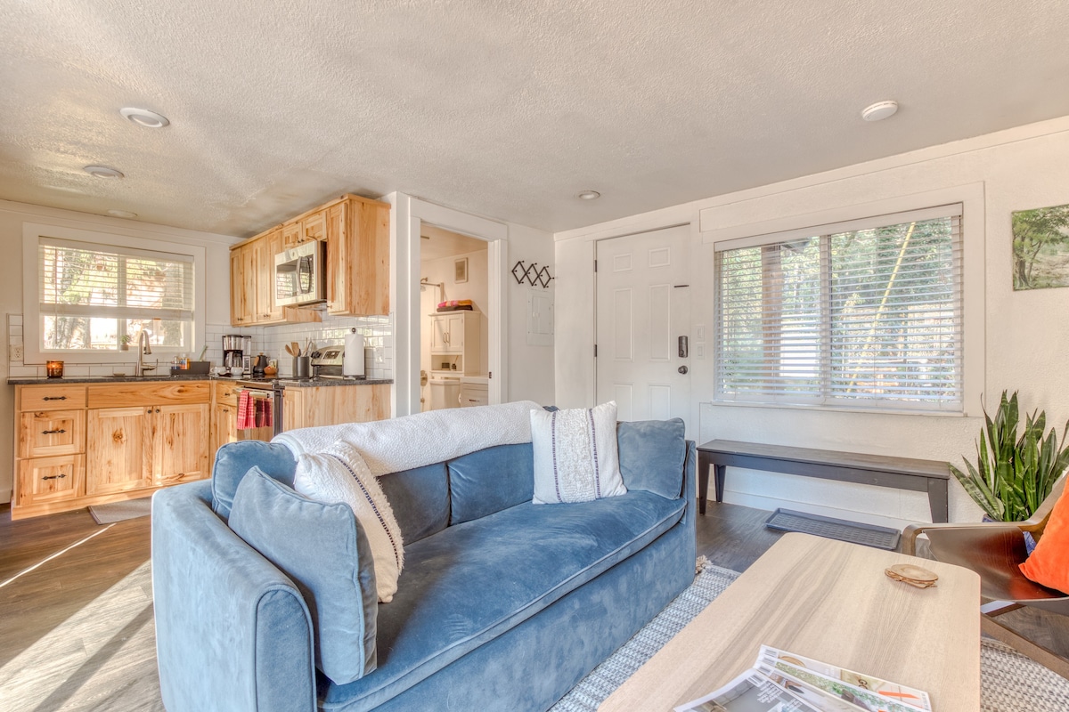 Cozy living room featuring a plush blue velvet sofa with decorative pillows, a light wood coffee table, and dark wood flooring, with a window offering natural light. The kitchen is visible in the background.