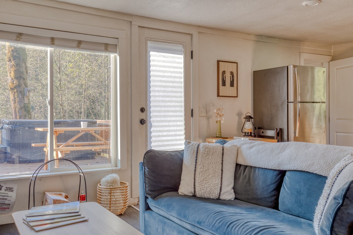 View from behind the light blue sofa, looking towards a large window and a glass door that opens to an outdoor deck with a hot tub and picnic table. A small wooden table with a lantern and framed art is visible next to the refrigerator.