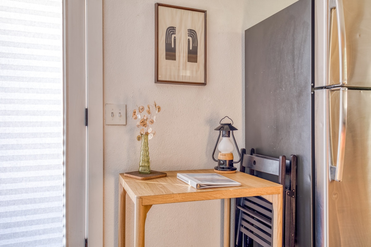 A cozy nook featuring a light wood table with a book, a vintage lantern, and a vase with dried flowers, positioned next to a dark refrigerator and a white door.