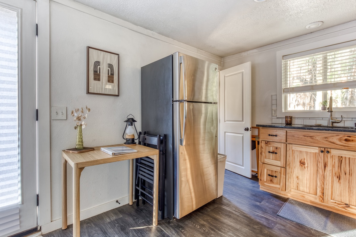 View of the kitchen area with light-colored wooden cabinets, dark countertops, and a stainless steel refrigerator. A window with white blinds is above the sink, and a small wooden table with a lantern is next to the refrigerator.