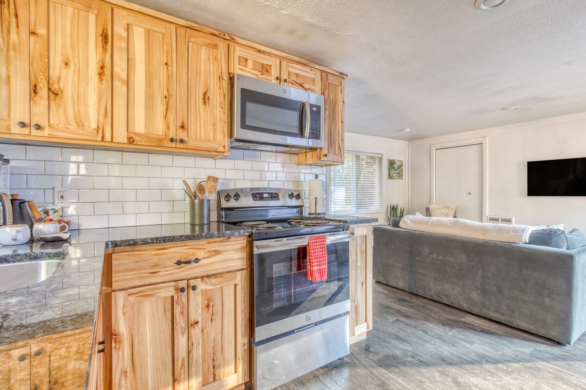 View of the kitchen with light wood cabinets, dark granite countertops, and stainless steel appliances, looking towards the living area with a gray sofa and a wall-mounted TV.