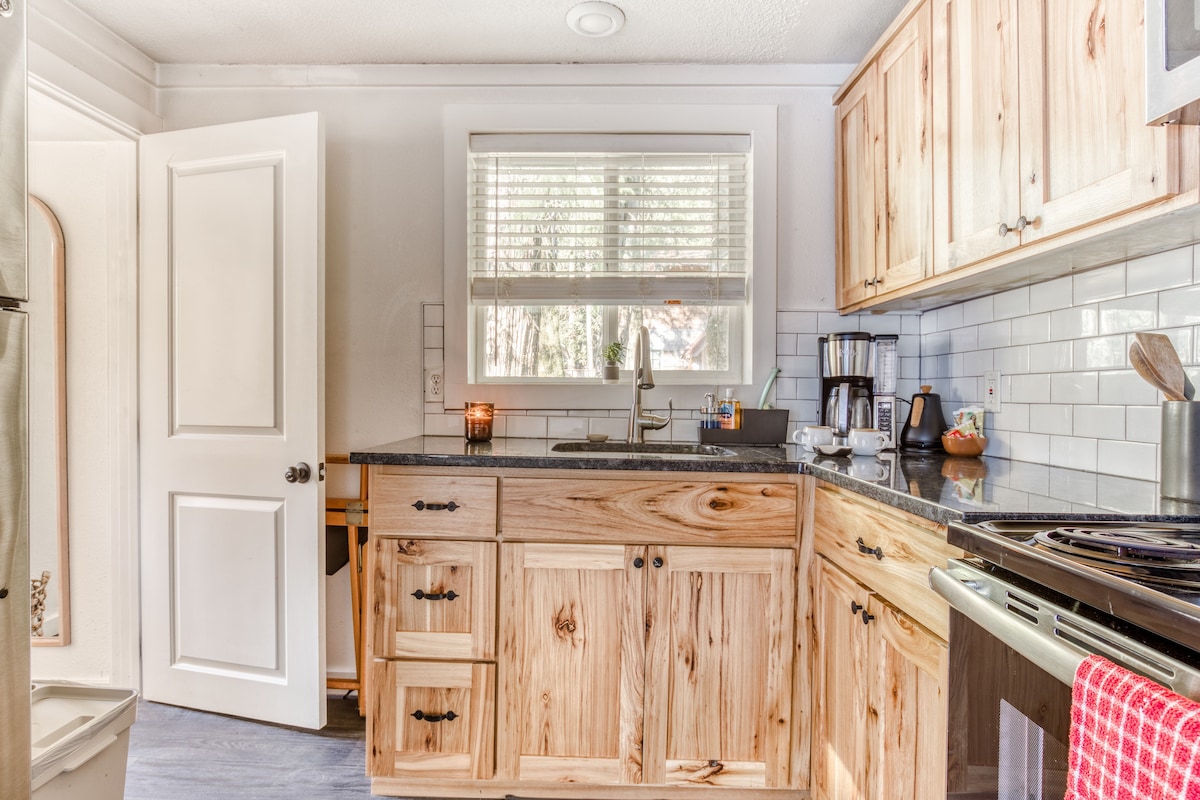 Corner view of a rustic kitchen with light wood cabinets, dark granite countertops, a white subway tile backsplash, and a window above the sink providing natural light.
