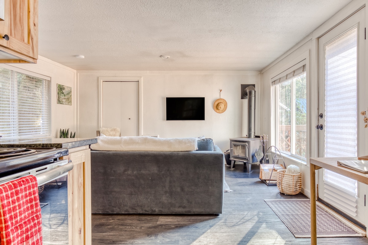 Open concept living area with a blue sofa facing a wood-burning stove, connected to the kitchen, with a large window and glass door leading to the outdoor deck.