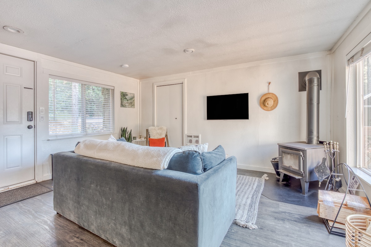 Bright living room with a large blue sofa, a wall-mounted TV, and a wood-burning stove, featuring a white entrance door and windows with blinds.