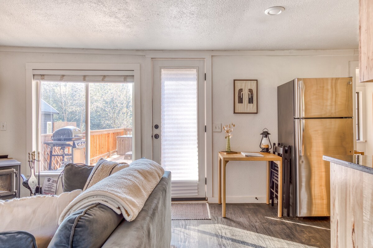 Angled shot of the living room showing the light blue sofa, a large window, and a glass door leading to the outdoor deck. A small wooden table with a lantern and framed art is positioned next to a stainless steel refrigerator, with the kitchen visible beyond.