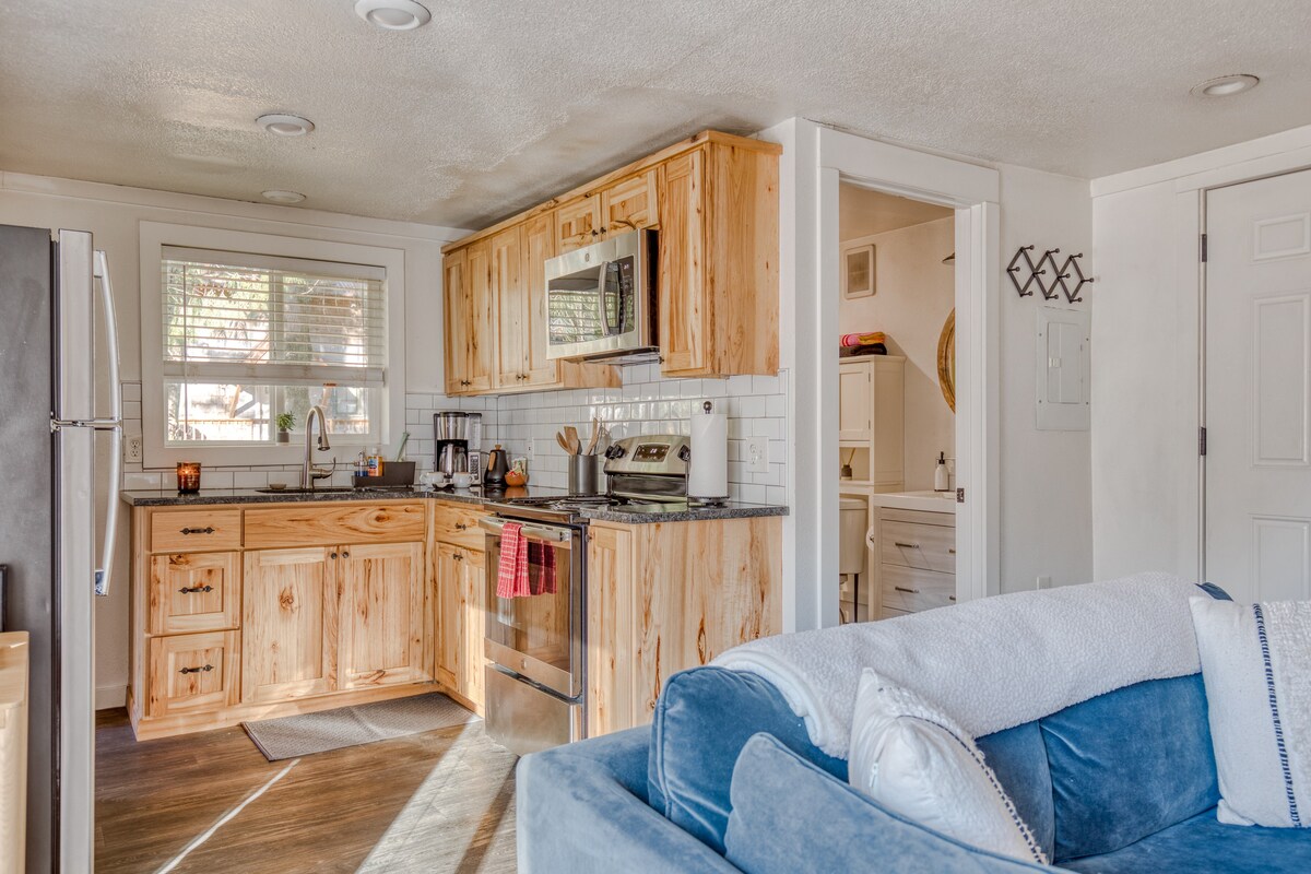 Open concept view showing the kitchen with light wood cabinets and dark countertops, flowing into a living area with a blue velvet sofa and dark wood flooring. A refrigerator is visible on the left.