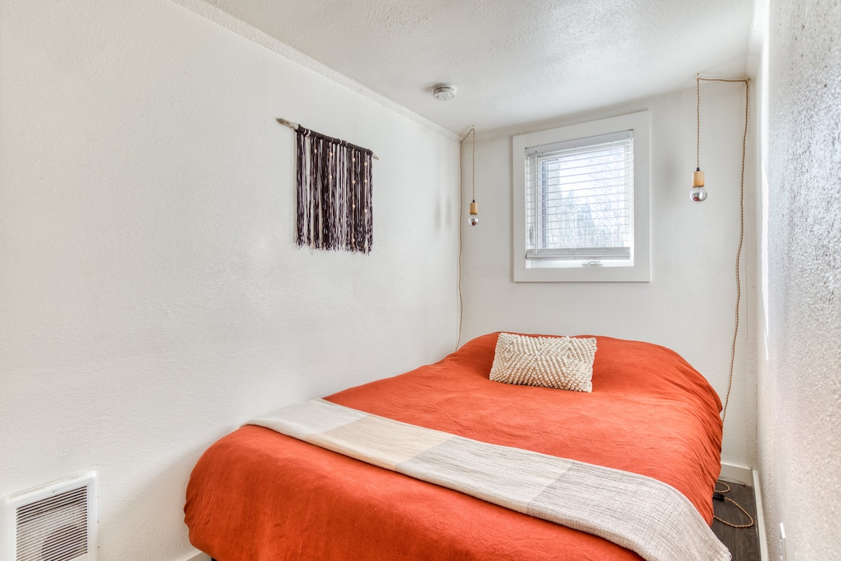 A cozy bedroom with a vibrant orange bedspread and a textured white throw pillow. A small window with blinds and a dark macrame wall hanging are visible on the white walls.