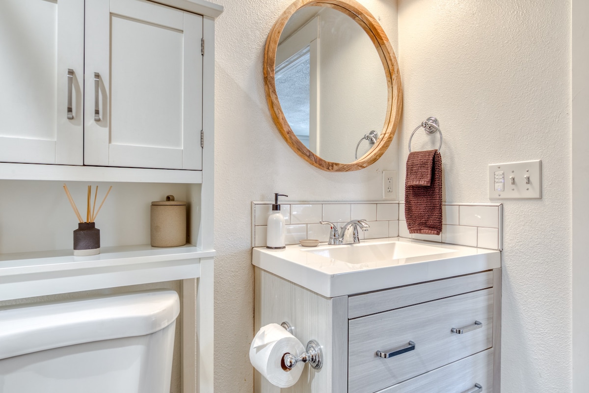A bright bathroom featuring a white vanity with a square sink, a round wooden-framed mirror, and a white cabinet above a toilet. A dark brown towel hangs on a ring.