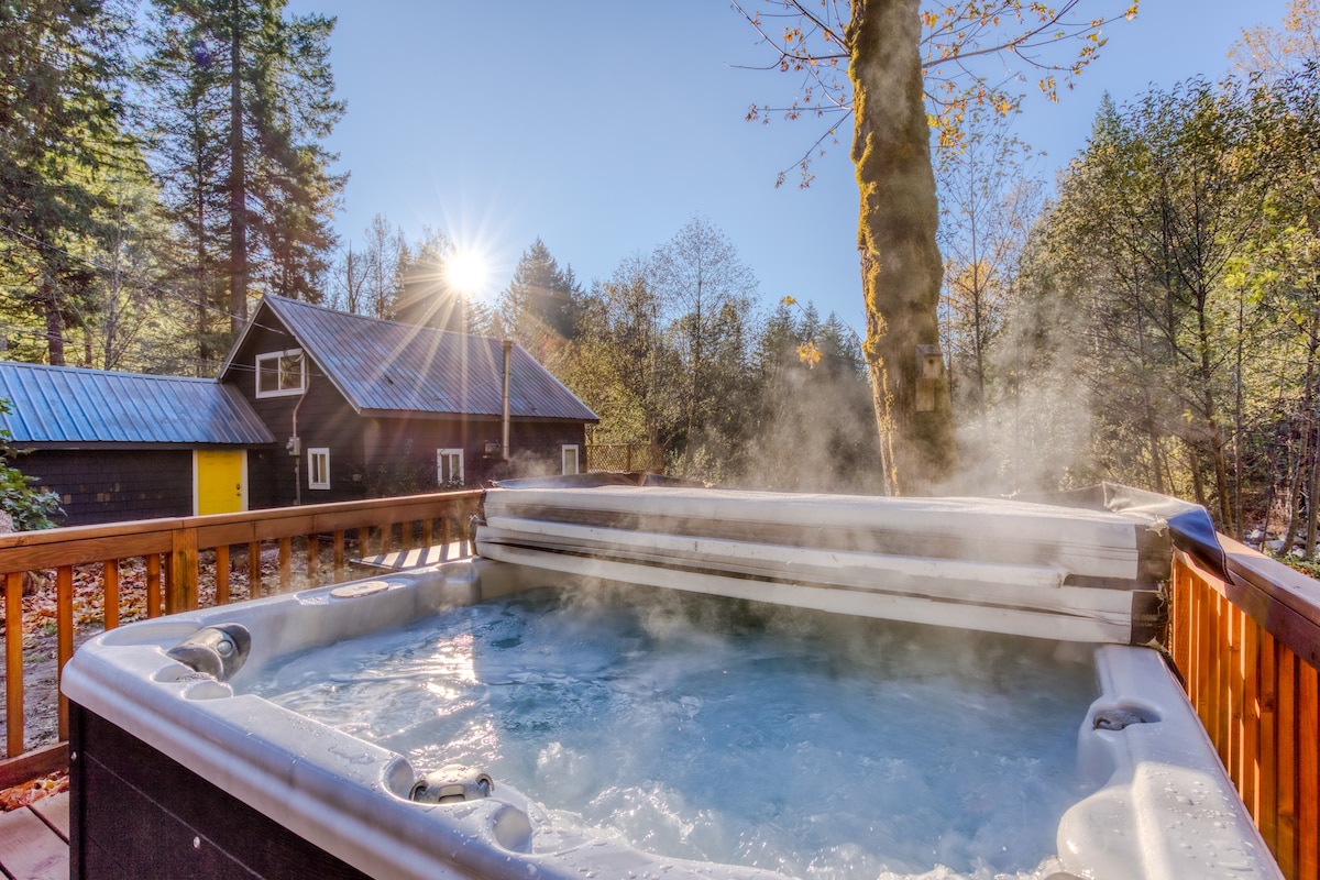 A steaming hot tub on a wooden deck, surrounded by lush green and autumn-colored trees under a bright blue sky. Sun rays peek through the trees, illuminating a dark cabin in the background.