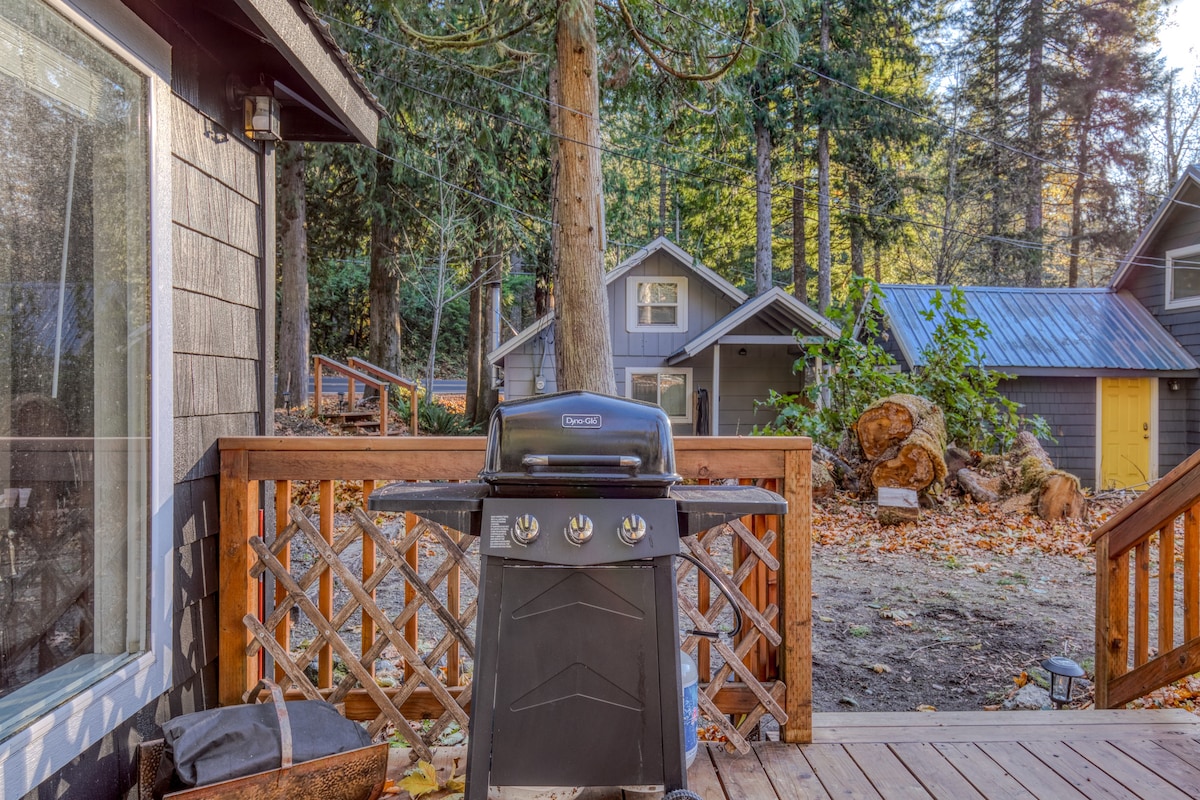 A black gas BBQ grill positioned on a wooden deck with a lattice railing. In the background, other cabins and tall trees are visible.