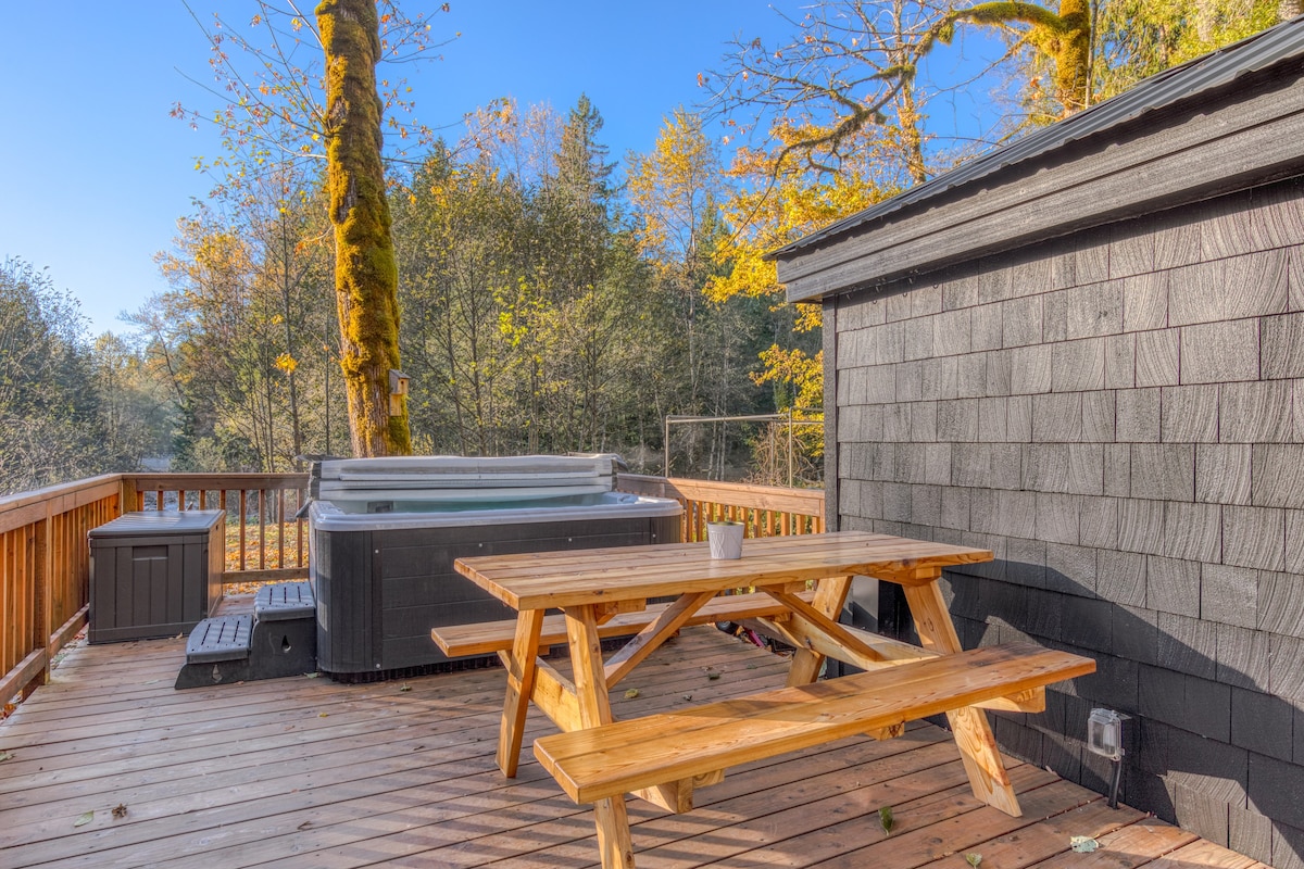 A spacious wooden deck featuring a rustic wooden picnic table and a dark hot tub with a closed cover. The deck overlooks a wooded area with autumn foliage.
