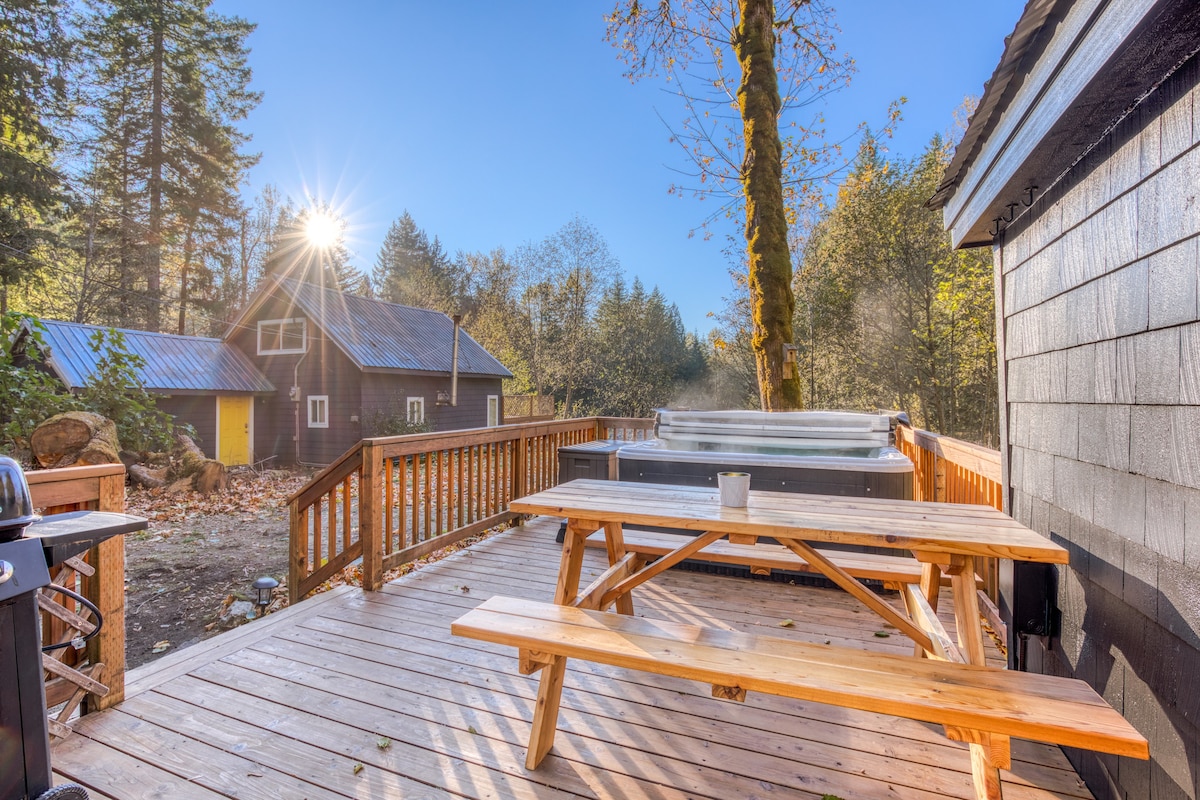 An expansive wooden deck with a picnic table and a hot tub, set against a backdrop of vibrant autumn trees and a dark cabin. Bright sunlight streams through the leaves, creating a warm glow.