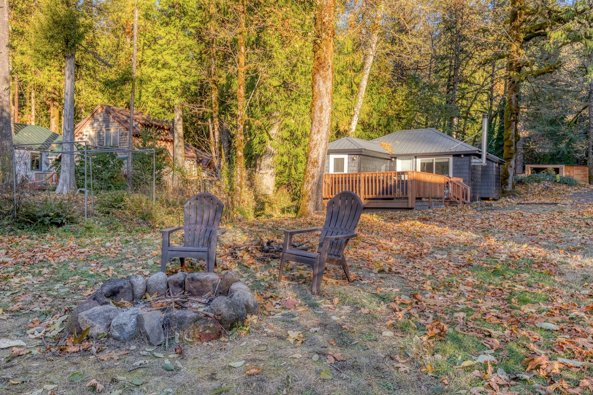 A stone fire pit surrounded by two dark Adirondack chairs in a grassy yard covered with fallen autumn leaves. A cabin and tall trees are in the background.
