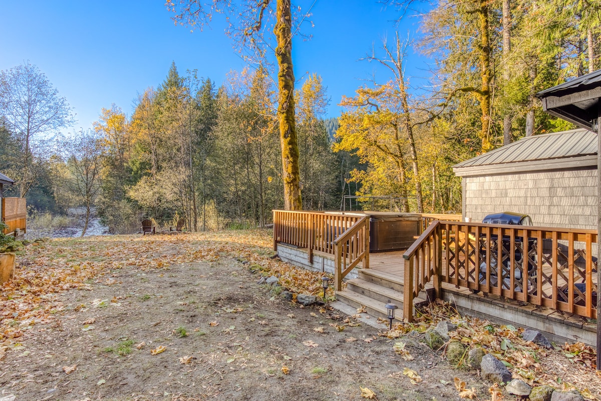 A wide view of a wooden deck with a hot tub, leading to a grassy yard scattered with autumn leaves. A river is visible in the distance, surrounded by dense trees under a clear blue sky.