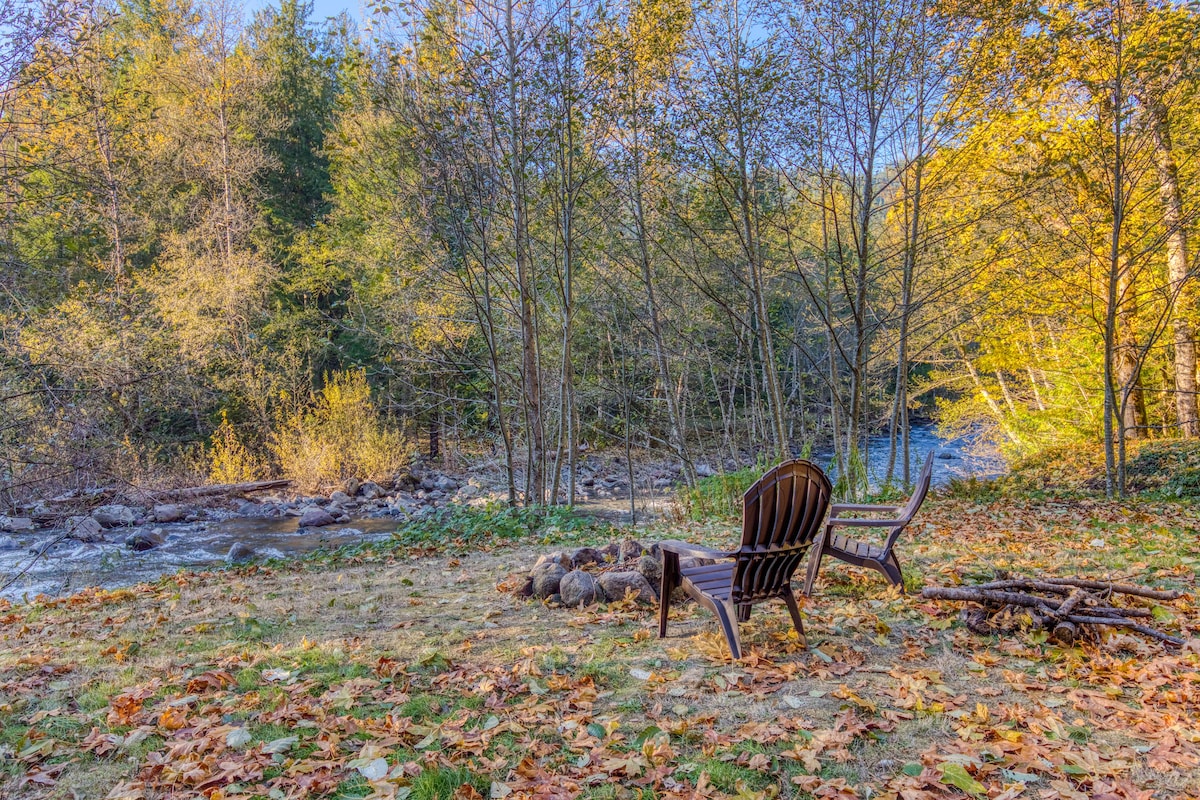 A rustic fire pit area with two Adirondack chairs, situated on a grassy bank next to a flowing river. The scene is framed by tall, slender trees with autumn foliage.