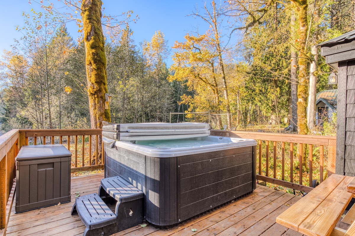 Large, dark-colored hot tub with steps sits on a spacious wooden deck, enclosed by a wooden railing, with a backdrop of vibrant autumn trees under a sunny sky.