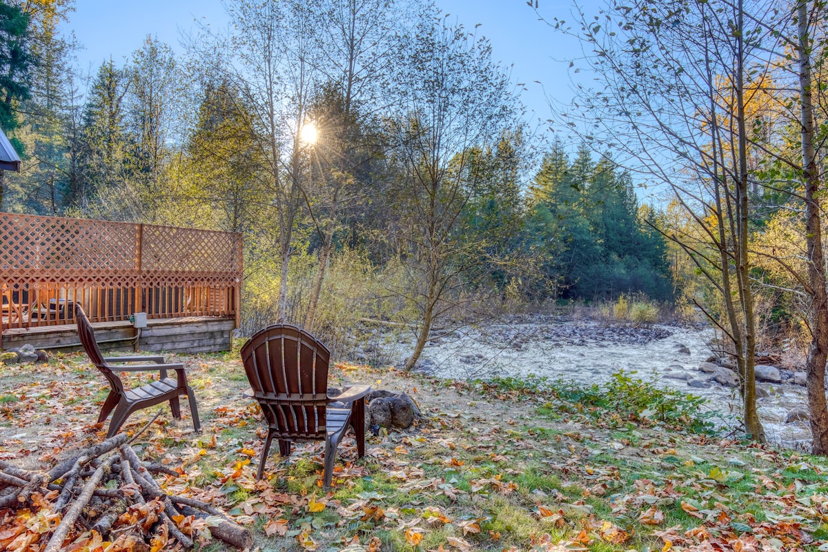 A sunny outdoor scene featuring a wooden deck with lattice railing, two Adirondack-style chairs, and a fire pit area with stacked logs, all overlooking a flowing river surrounded by lush trees.