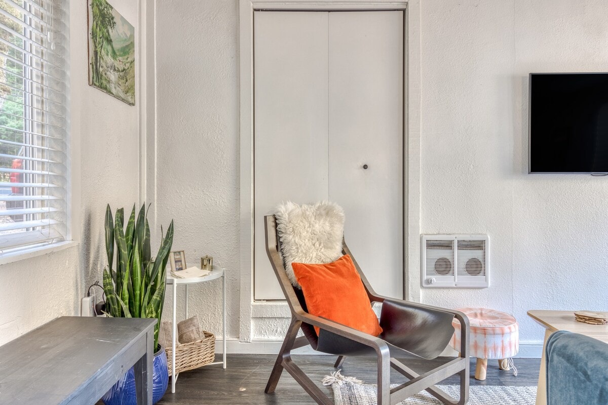 Close-up of a cozy corner in the living room featuring a modern wooden armchair with a black leather sling, an orange throw pillow, and a white faux fur accent. A snake plant in a blue pot sits on a dark bench next to a window with white blinds.