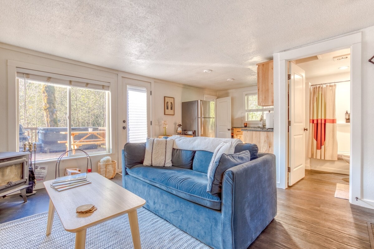 View from behind the light blue velvet sofa, looking towards the kitchen and bathroom. A large window and a glass door provide natural light, revealing a glimpse of the outdoor deck and a hot tub.