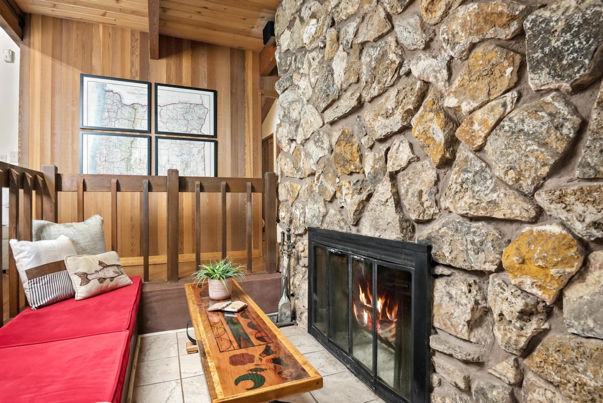 A cozy nook with a large stone fireplace, a built-in red cushioned bench, and a unique wooden coffee table, featuring wood-paneled walls and a view into the main living area.