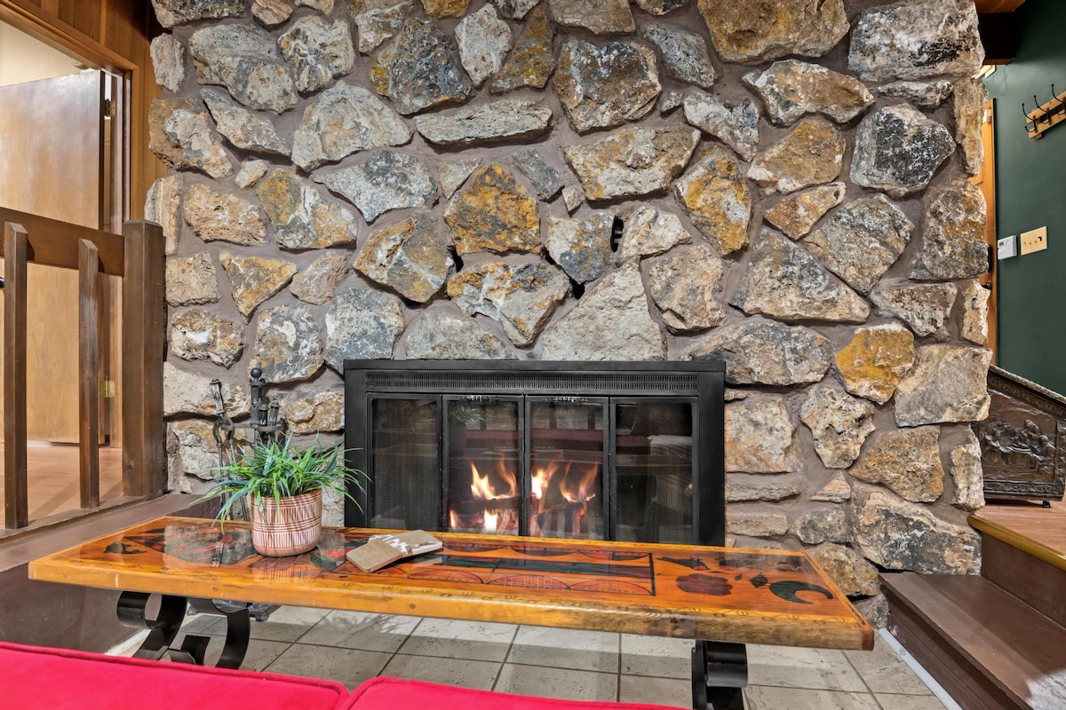 A close-up of a rustic stone fireplace with a roaring fire behind a glass screen, flanked by a red cushioned bench and a unique wooden coffee table with carved details.