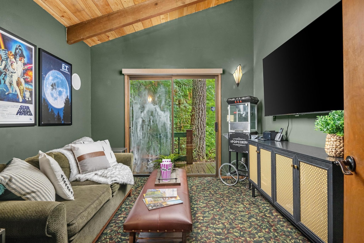 A cozy media room with a green sofa, a leather ottoman coffee table, and a large wall-mounted TV, featuring a popcorn machine and movie posters, with a sliding door leading to an outdoor deck.