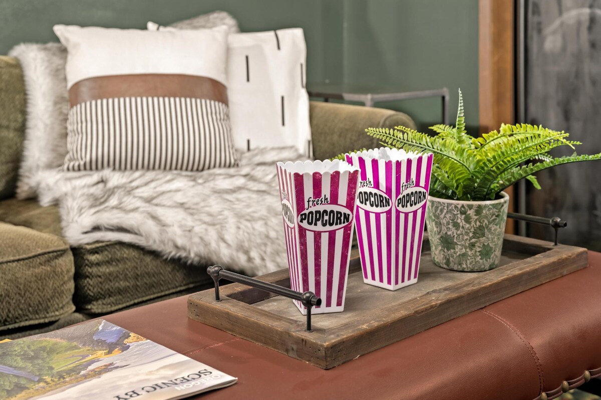 Close-up of a brown leather ottoman serving as a coffee table, holding a wooden tray with two striped popcorn containers and a potted fern, with a green sofa and decorative pillows in the background.