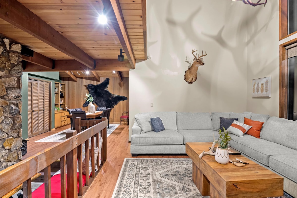 Spacious living room with a large light gray sectional sofa, a rustic wooden coffee table, and a patterned area rug, featuring a deer head mount on a light-colored wall and a stone fireplace on the left.