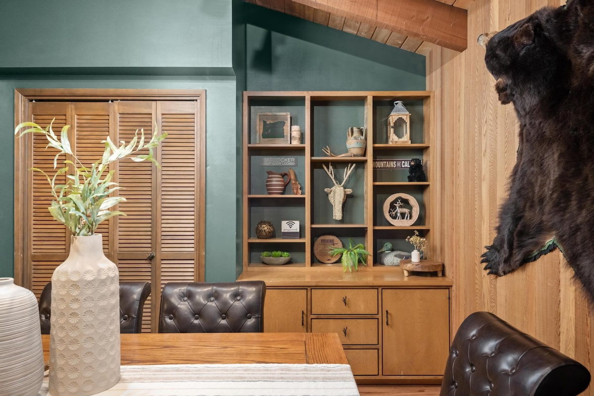 Dining area with a wooden table and dark leather chairs, adjacent to a built-in wooden shelving unit displaying various decorative items, and a large bear rug hanging on a wood-paneled wall.