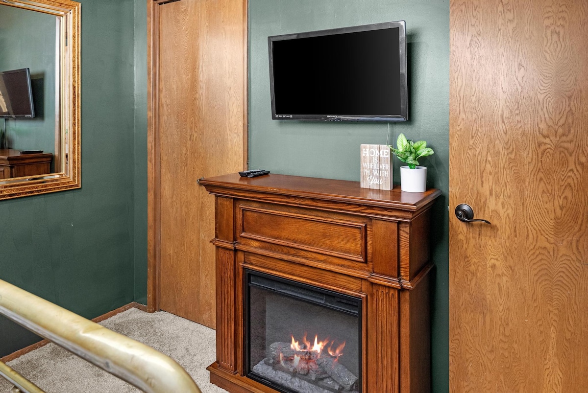 Cozy landing area featuring a wall-mounted TV above a wooden electric fireplace, with a small potted plant and a decorative sign, flanked by wooden doors and a gold-framed mirror.