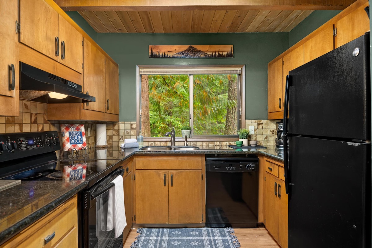 U-shaped kitchen with light wood cabinetry, black granite countertops, and black appliances, featuring a window above the sink offering a view of lush green trees, and a tiled backsplash.