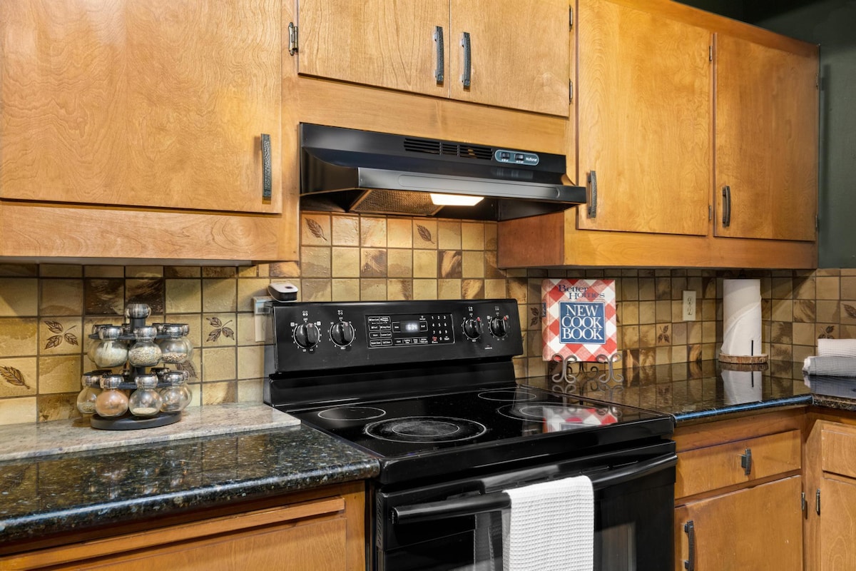 Close-up of the kitchen stove area, showing a black electric range with an overhead microwave/vent hood, black granite countertops, and a spice rack, with light wood cabinets above.
