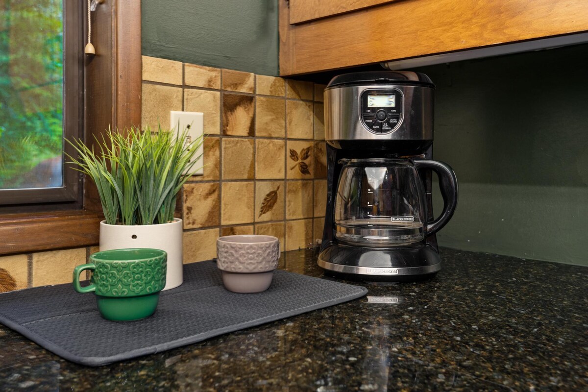 Corner of the kitchen countertop featuring a black coffee maker, two ceramic mugs, and a small potted plant, set against a tiled backsplash and a window.