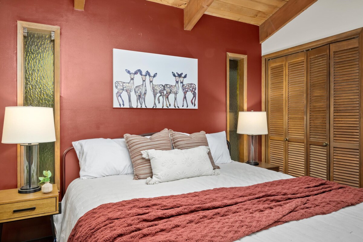 Bedroom with a queen-sized bed dressed in white linens and a rust-colored throw, flanked by wooden nightstands with lamps, and a large deer-themed artwork on a dark red accent wall.