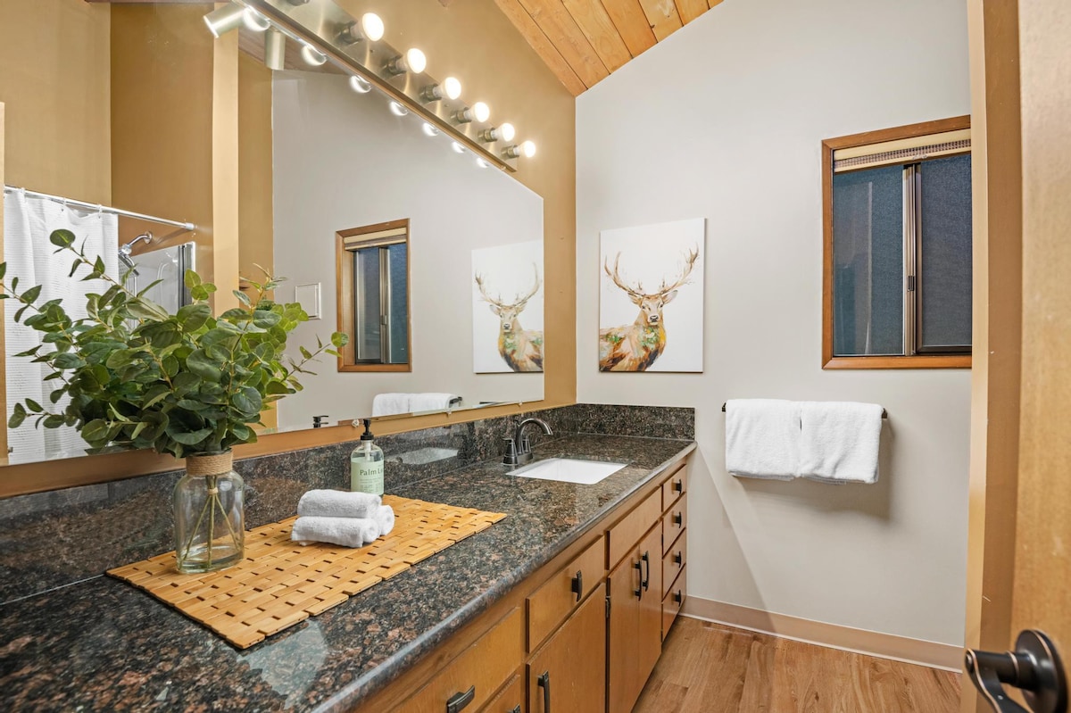 Bathroom with a long single vanity featuring a white sink set into a dark granite countertop, a large mirror with Hollywood-style lighting, and deer-themed wall art, with a shower/tub combo visible.