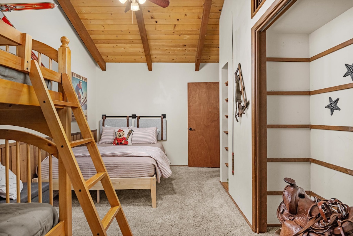 A cozy bedroom featuring a wooden bunk bed on the left and a queen bed with striped bedding on the right, all under a vaulted wooden ceiling with exposed beams. A built-in shelving unit with western decor is visible.